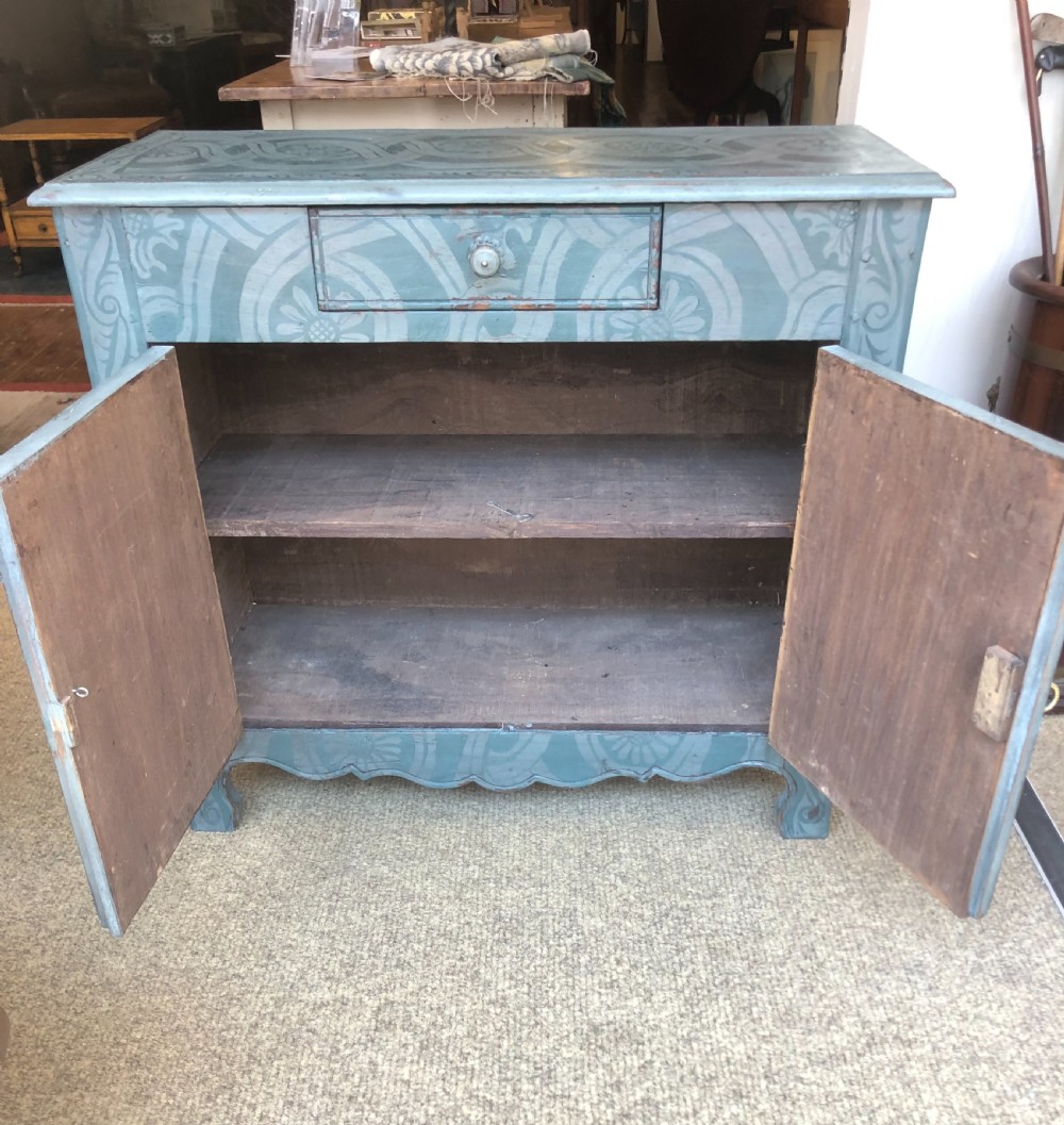 A small wooden cupboard hand-painted in muted sage green with light grey strapwork and floral motifs, featuring a single top drawer and two panelled doors. View V