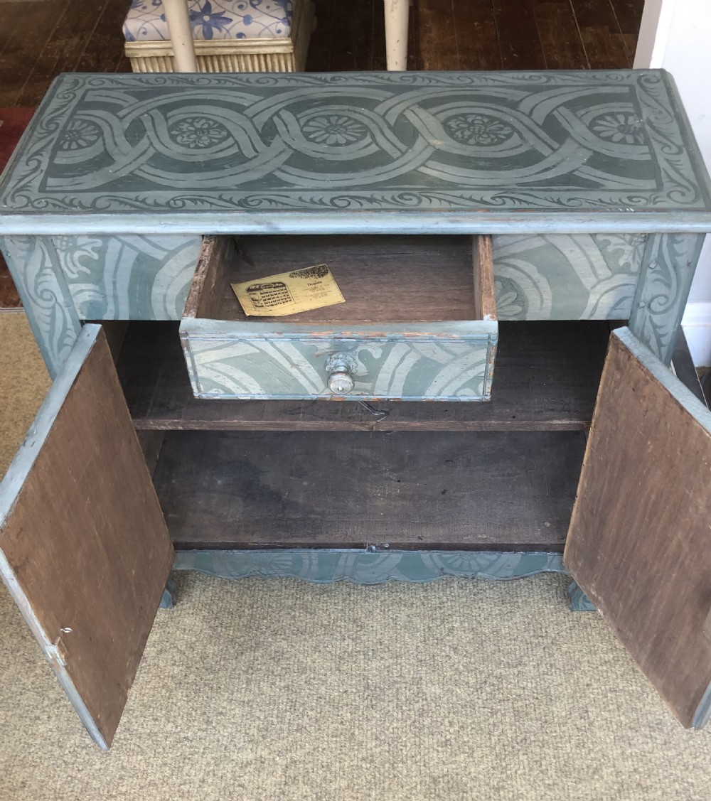 A small wooden cupboard hand-painted in muted sage green with light grey strapwork and floral motifs, featuring a single top drawer and two panelled doors. View VII
