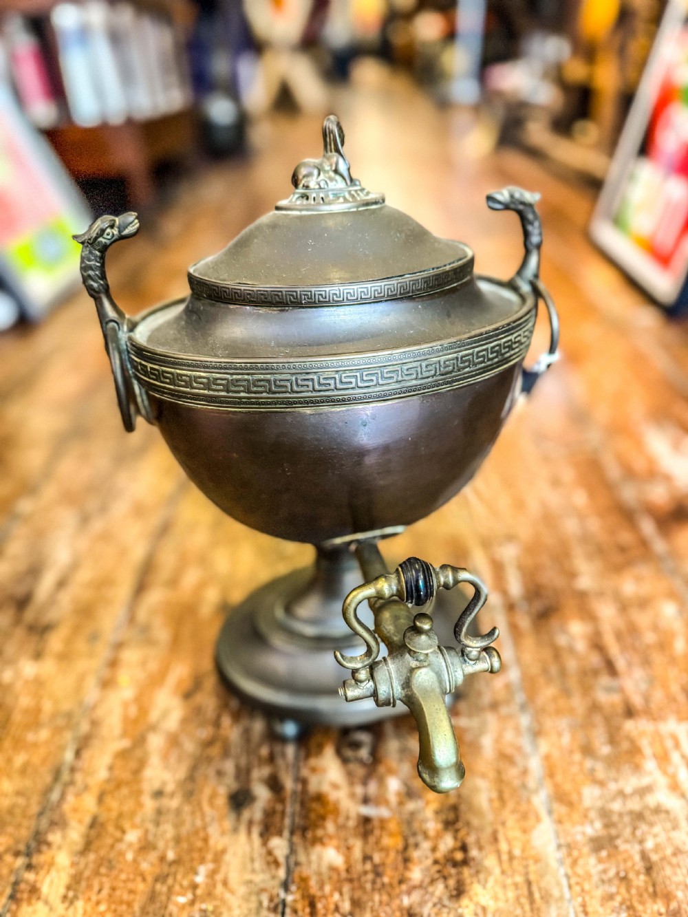 An antique Regency metal tea urn featuring a dome lid with finial, Greek key engraved band, zoomorphic side handles, and a brass spigot on a pedestal base.