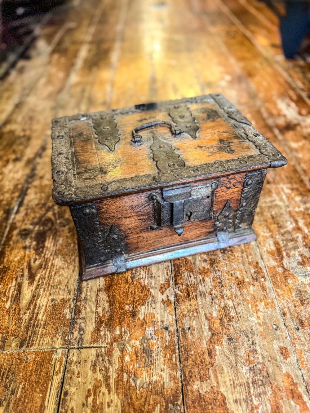 A 17th-century rectangular wooden box featuring heavy wrought-iron corner braces, decorative straps, a top carrying handle, and an oversized front lockplate.