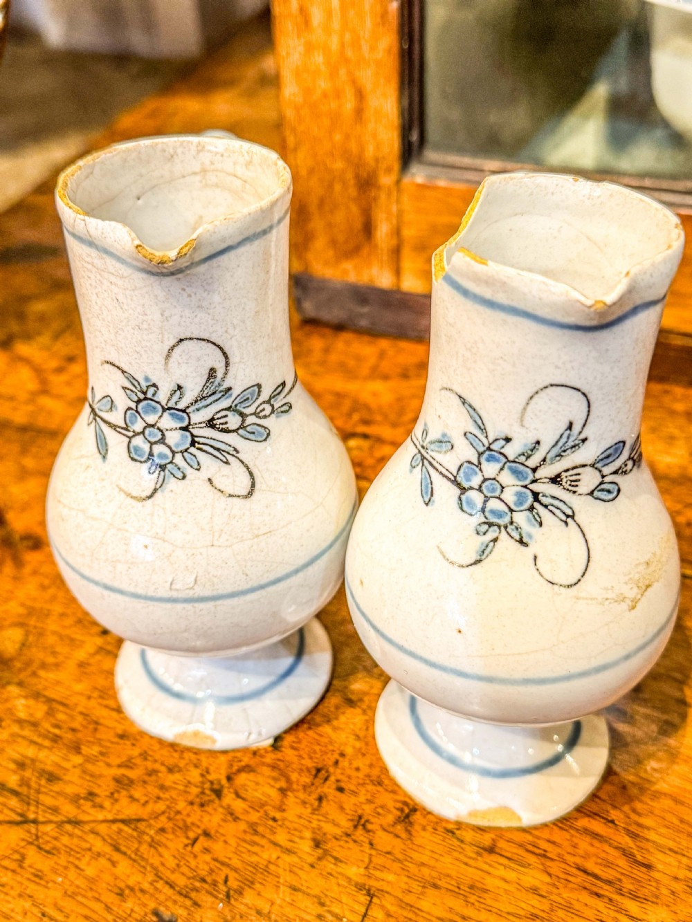 A pair of small antique off-white tin-glazed earthenware jugs decorated with hand-painted blue floral motifs on pedestal bases.