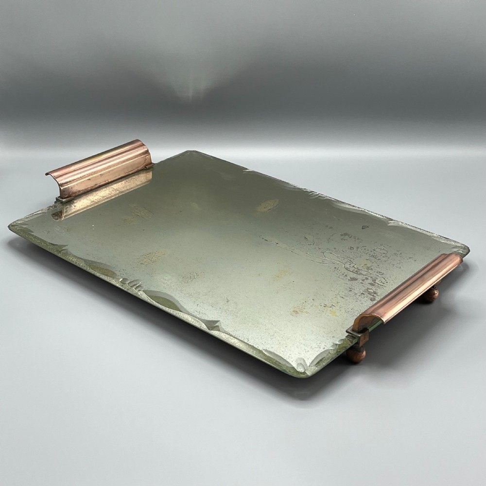 A rectangular Art Deco mirrored glass tray featuring cut decorative edges and rolled copper handles supported by small footings.