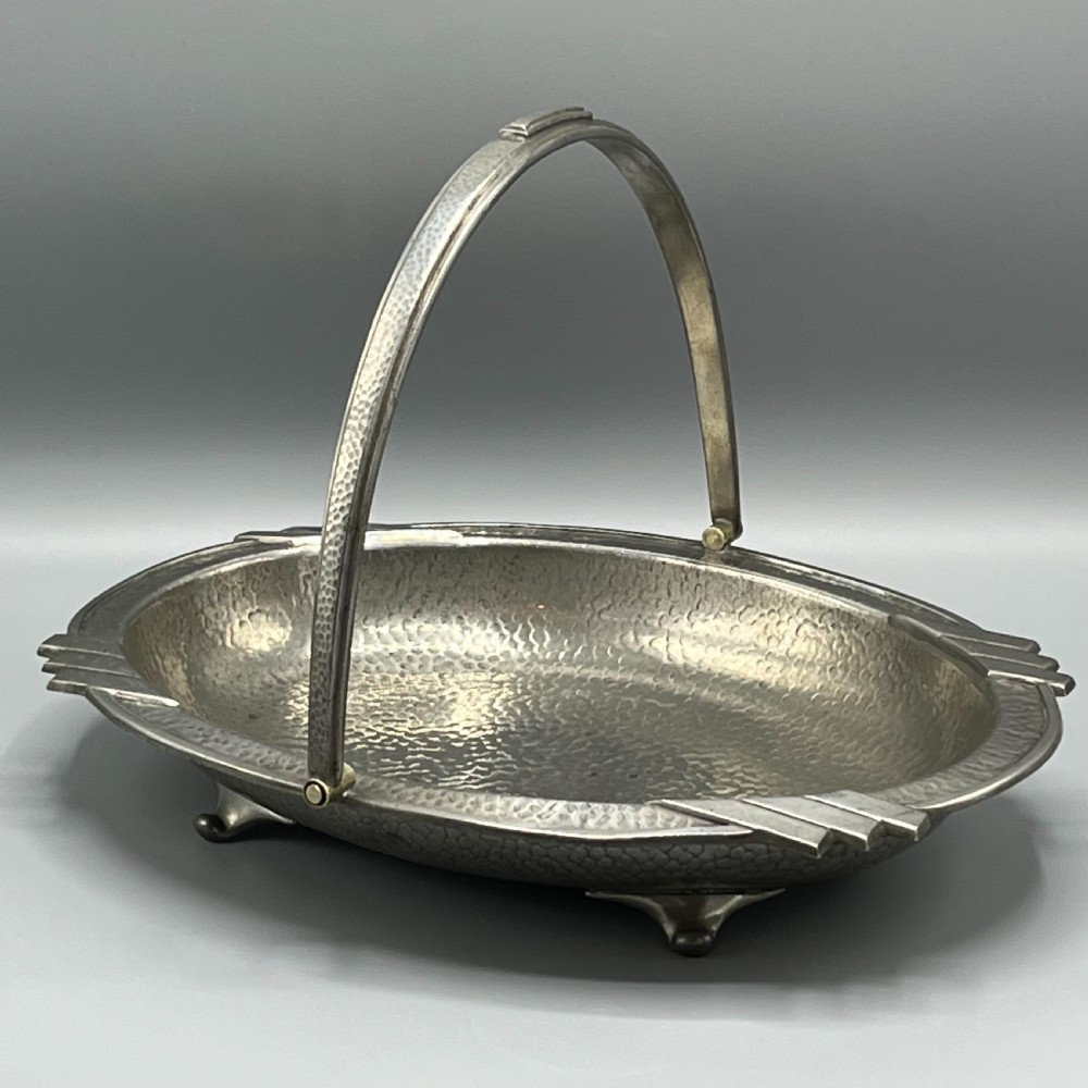 An oval hand-hammered pewter fruit basket featuring a high arching handle, geometric rim details, and small cast feet.