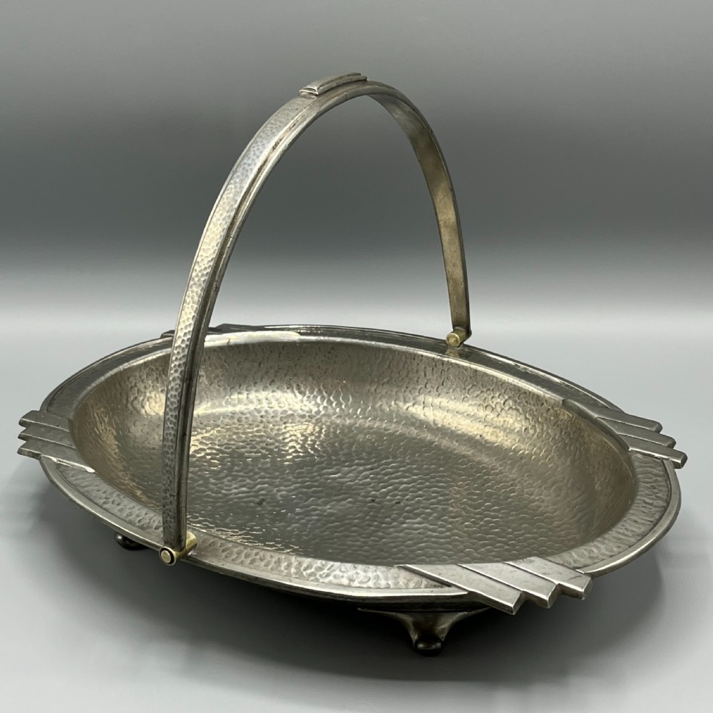 An oval hand-hammered pewter fruit basket featuring a high arching handle, geometric rim details, and small cast feet. View II