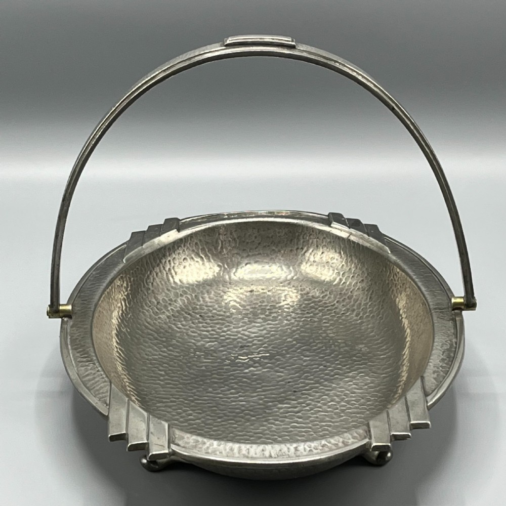 An oval hand-hammered pewter fruit basket featuring a high arching handle, geometric rim details, and small cast feet. View III