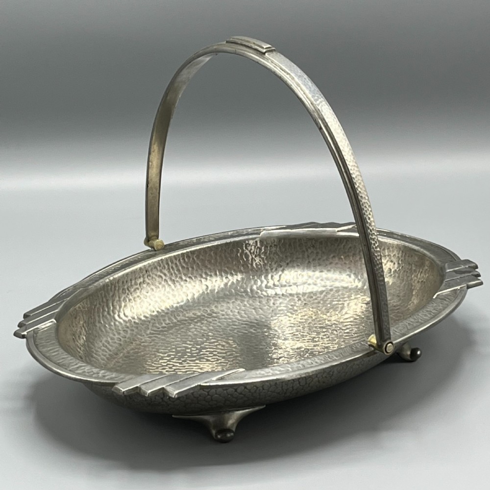 An oval hand-hammered pewter fruit basket featuring a high arching handle, geometric rim details, and small cast feet. View IV