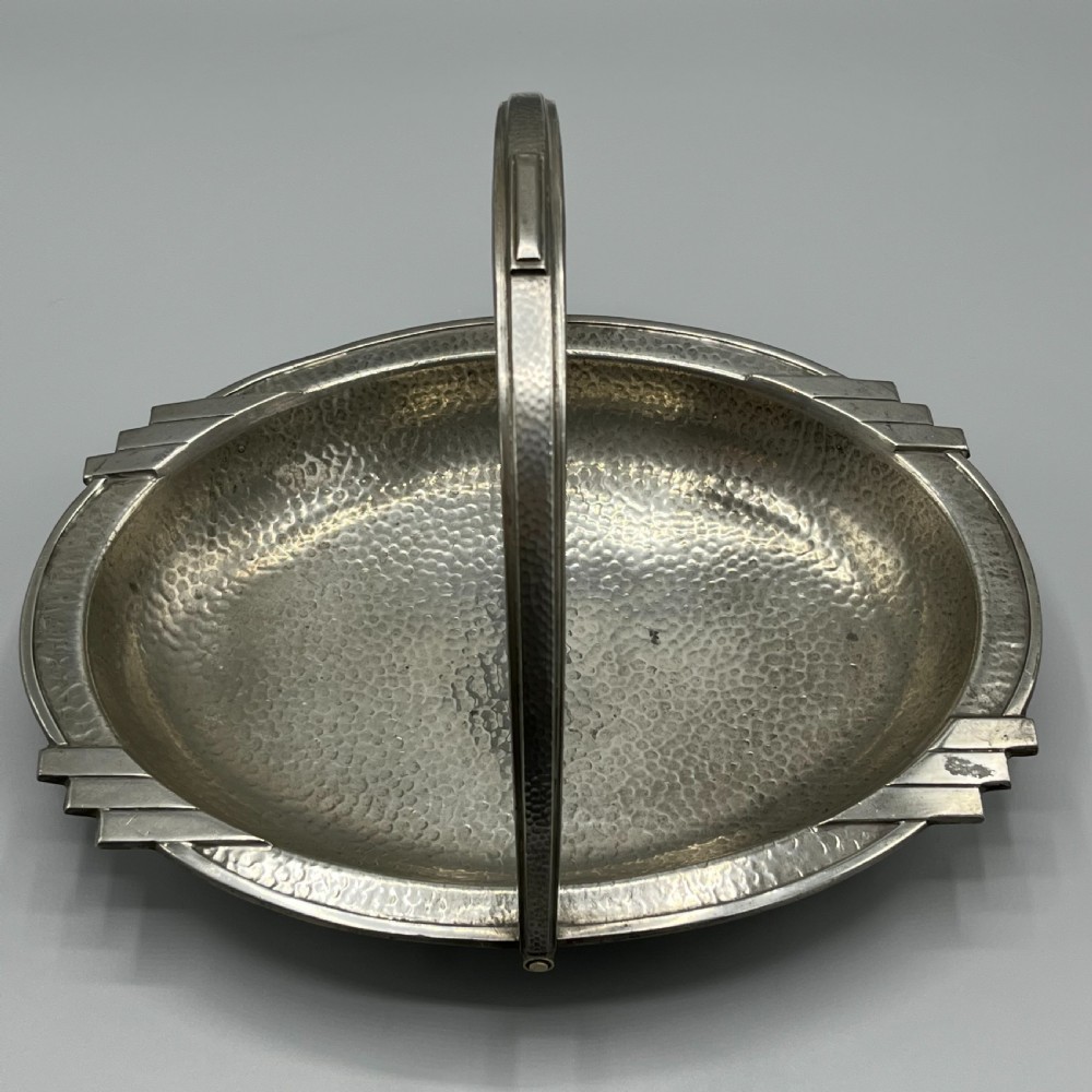 An oval hand-hammered pewter fruit basket featuring a high arching handle, geometric rim details, and small cast feet. View V
