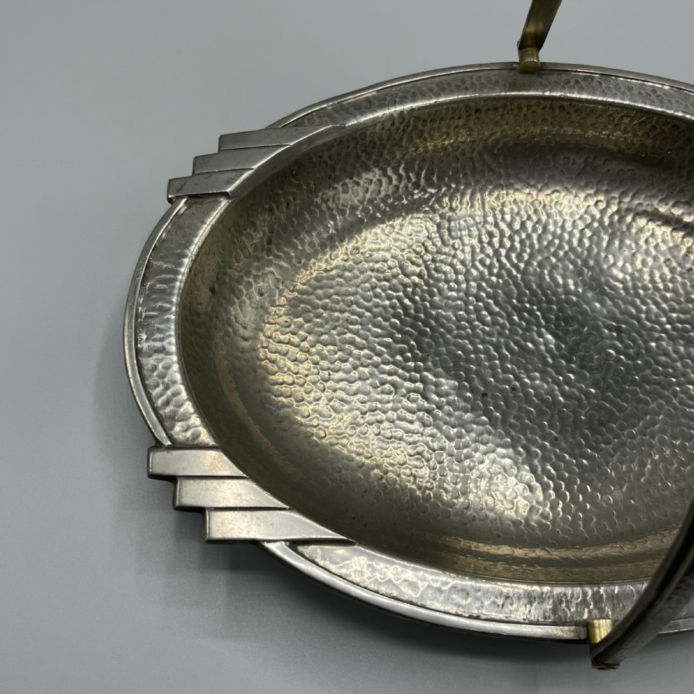 An oval hand-hammered pewter fruit basket featuring a high arching handle, geometric rim details, and small cast feet. View VI