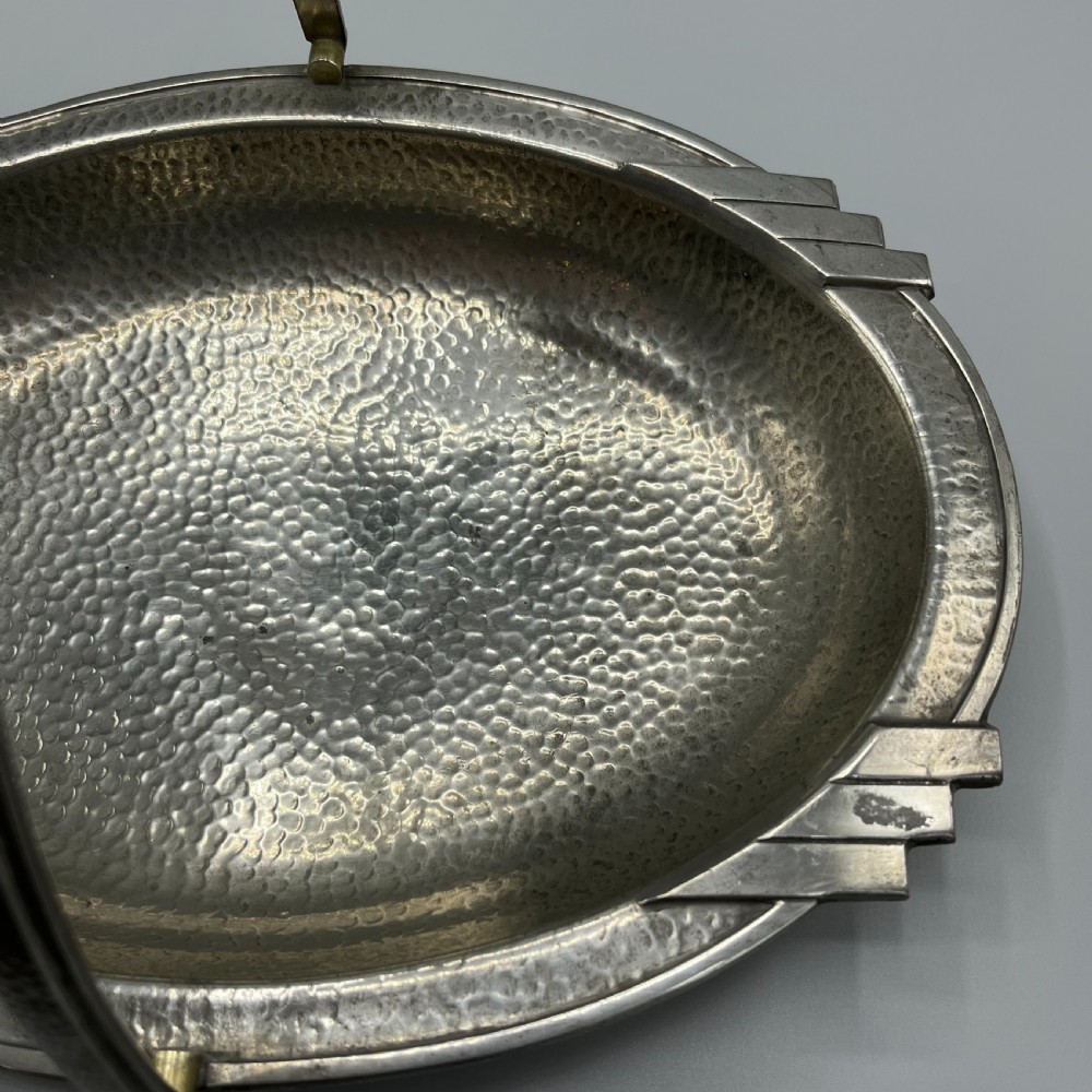 An oval hand-hammered pewter fruit basket featuring a high arching handle, geometric rim details, and small cast feet. View VII