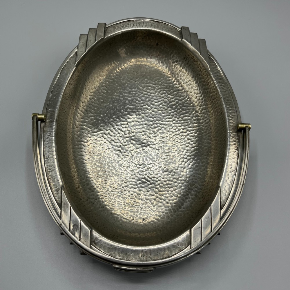 An oval hand-hammered pewter fruit basket featuring a high arching handle, geometric rim details, and small cast feet. View VIII
