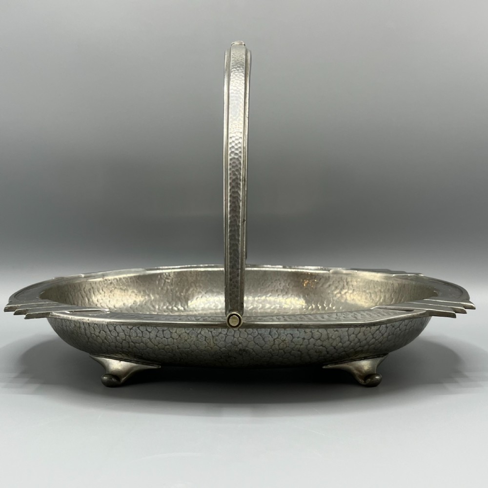 An oval hand-hammered pewter fruit basket featuring a high arching handle, geometric rim details, and small cast feet. View IX