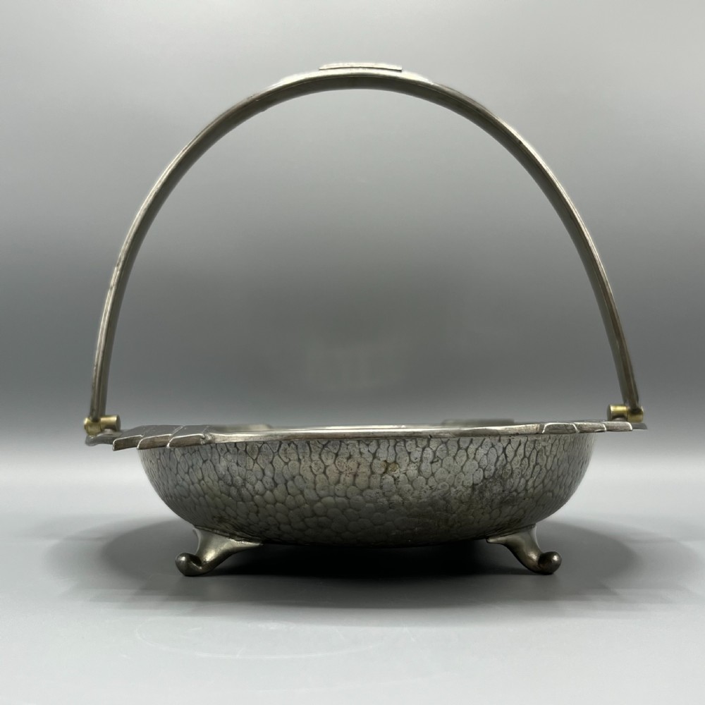 An oval hand-hammered pewter fruit basket featuring a high arching handle, geometric rim details, and small cast feet. View X