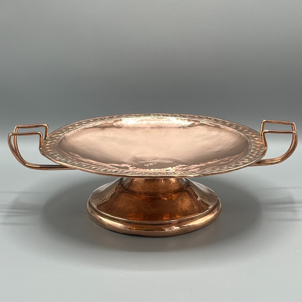 A hand-hammered copper tazza featuring twin side handles, a circular pedestal base, and a stamped geometric border along the shallow bowl rim.