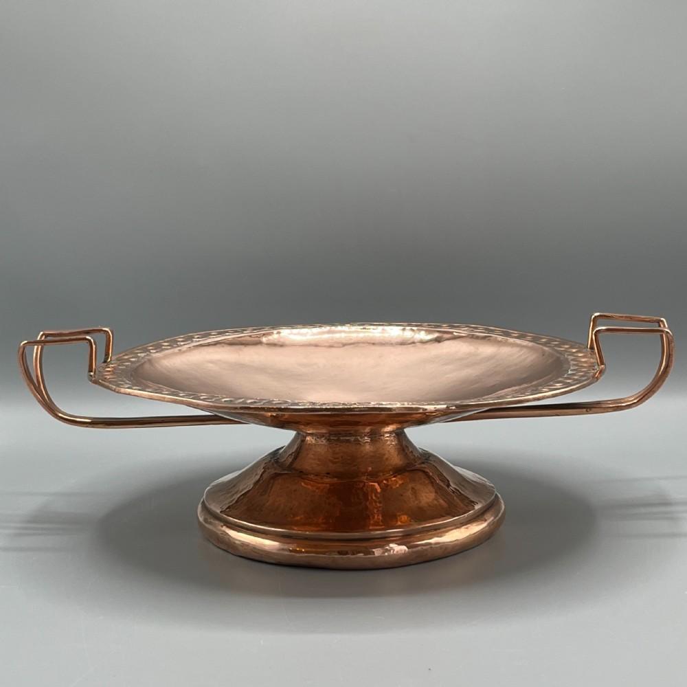 A hand-hammered copper tazza featuring twin side handles, a circular pedestal base, and a stamped geometric border along the shallow bowl rim. View II