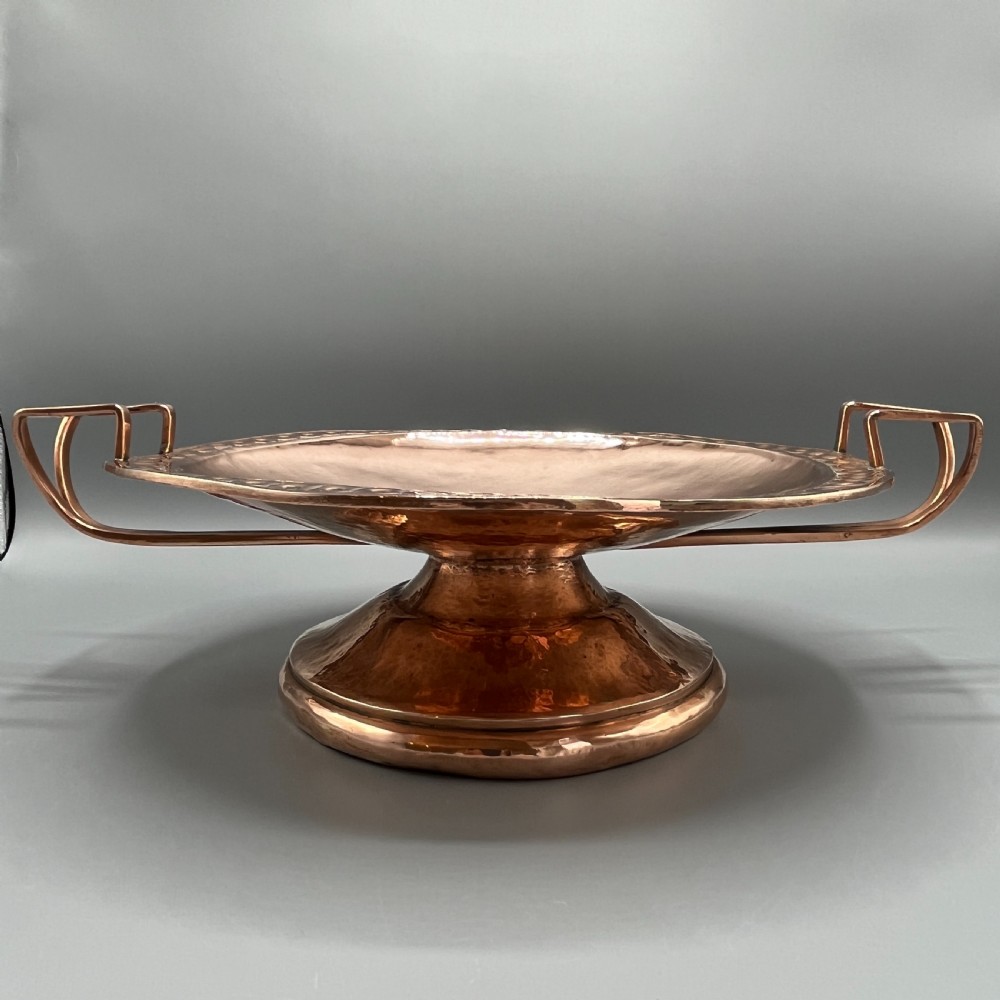 A hand-hammered copper tazza featuring twin side handles, a circular pedestal base, and a stamped geometric border along the shallow bowl rim. View III