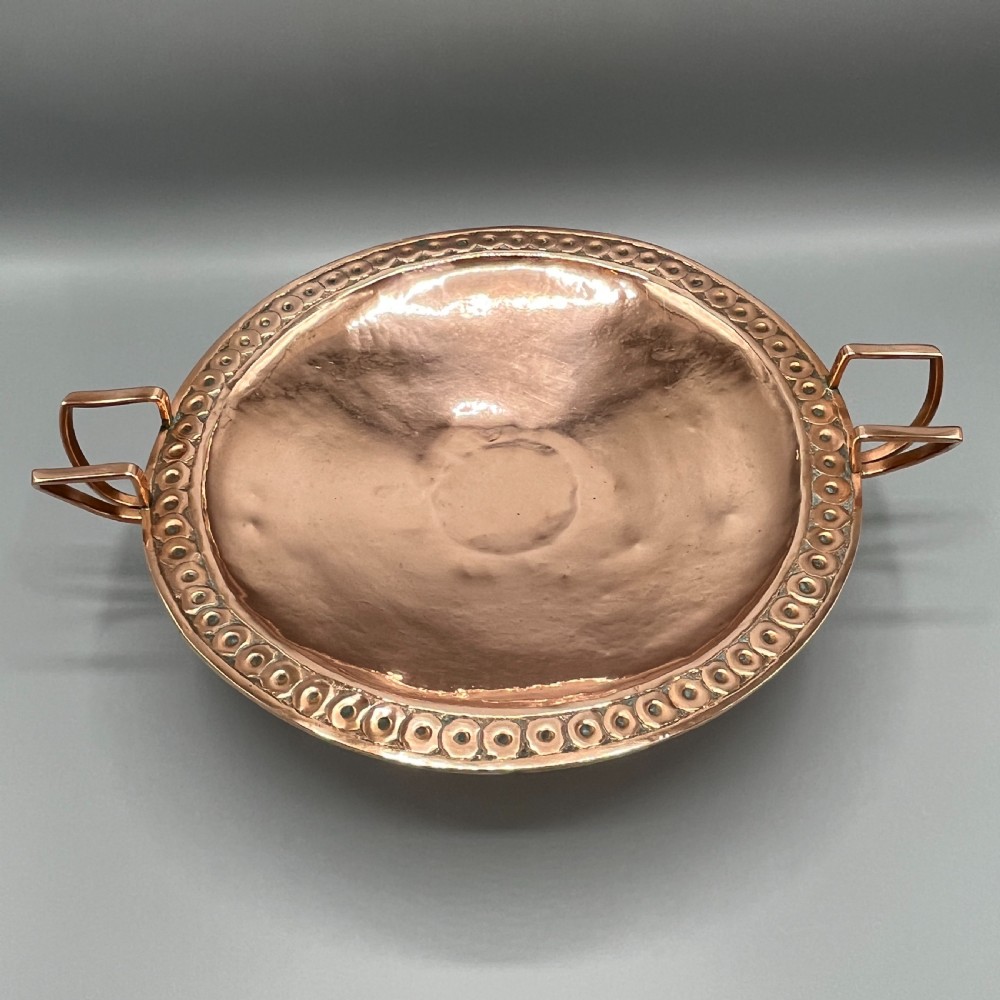A hand-hammered copper tazza featuring twin side handles, a circular pedestal base, and a stamped geometric border along the shallow bowl rim. View IV