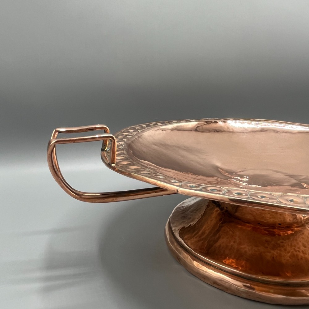 A hand-hammered copper tazza featuring twin side handles, a circular pedestal base, and a stamped geometric border along the shallow bowl rim. View V