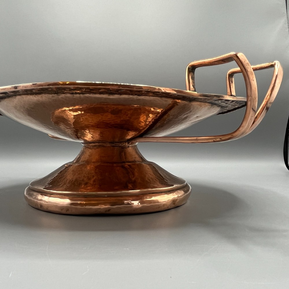 A hand-hammered copper tazza featuring twin side handles, a circular pedestal base, and a stamped geometric border along the shallow bowl rim. View VI