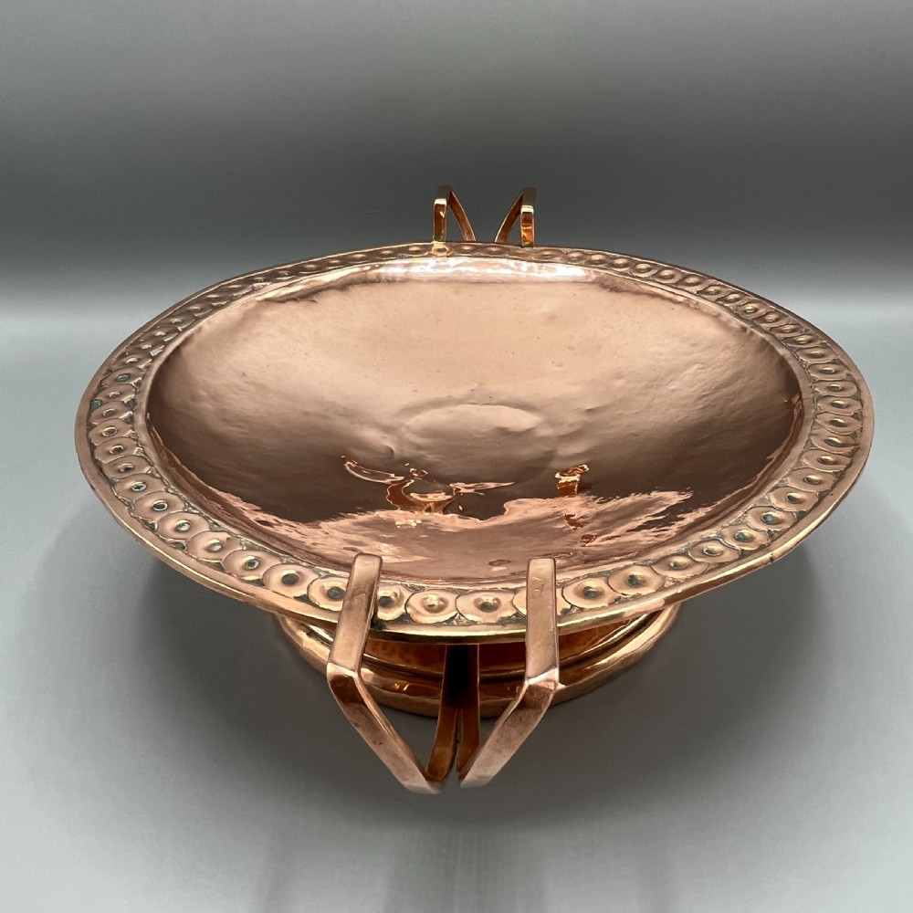 A hand-hammered copper tazza featuring twin side handles, a circular pedestal base, and a stamped geometric border along the shallow bowl rim. View VII