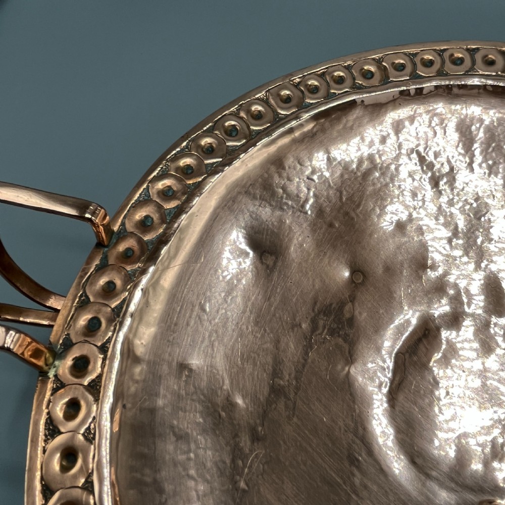 A hand-hammered copper tazza featuring twin side handles, a circular pedestal base, and a stamped geometric border along the shallow bowl rim. View X