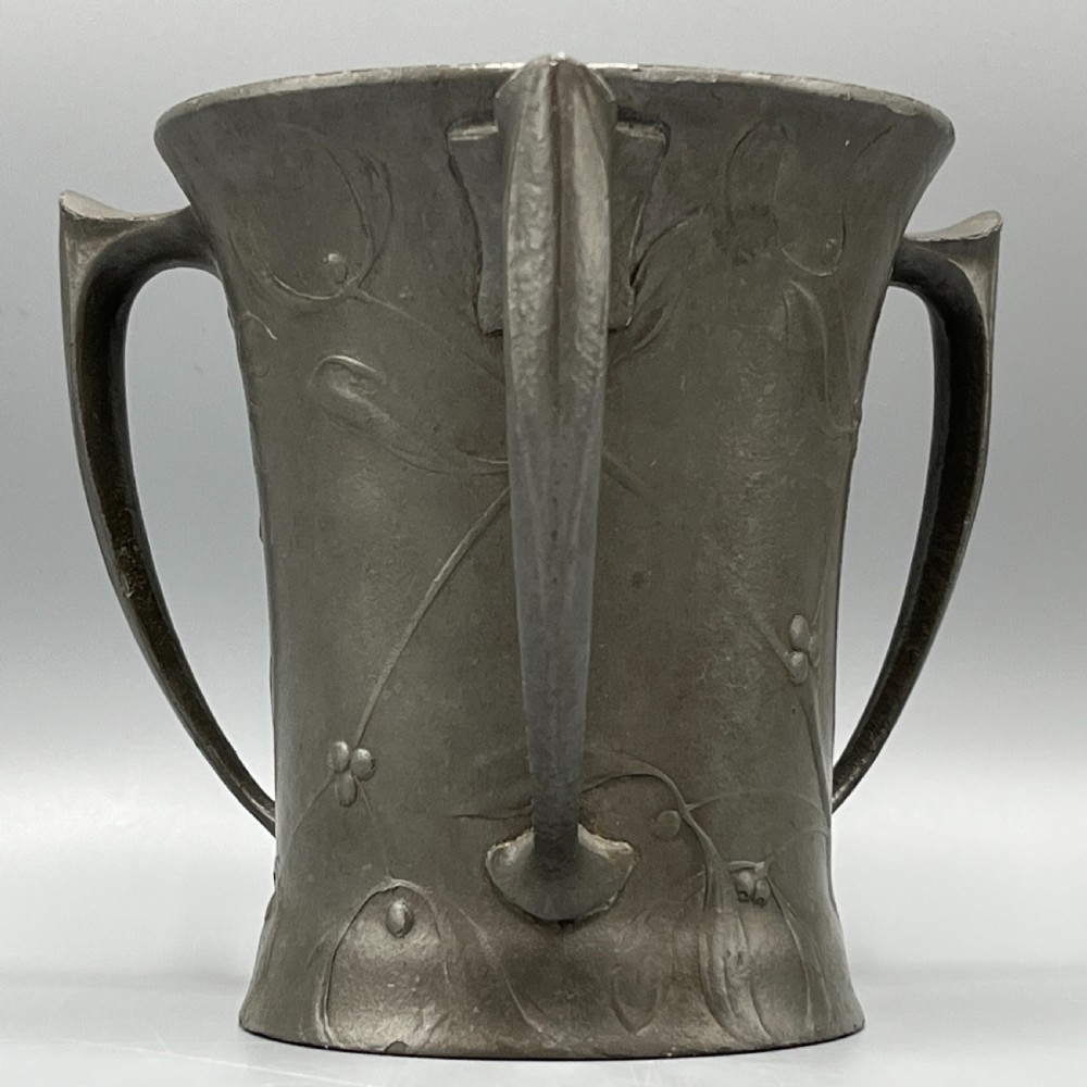 A grey pewter Art Nouveau tyg vase featuring three curved handles and relief-moulded mistletoe motifs around the body.