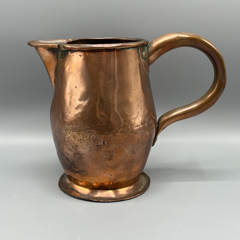 An antique Georgian copper jug featuring a hand-hammered body, wide pouring spout, flared base, and a riveted loop handle.