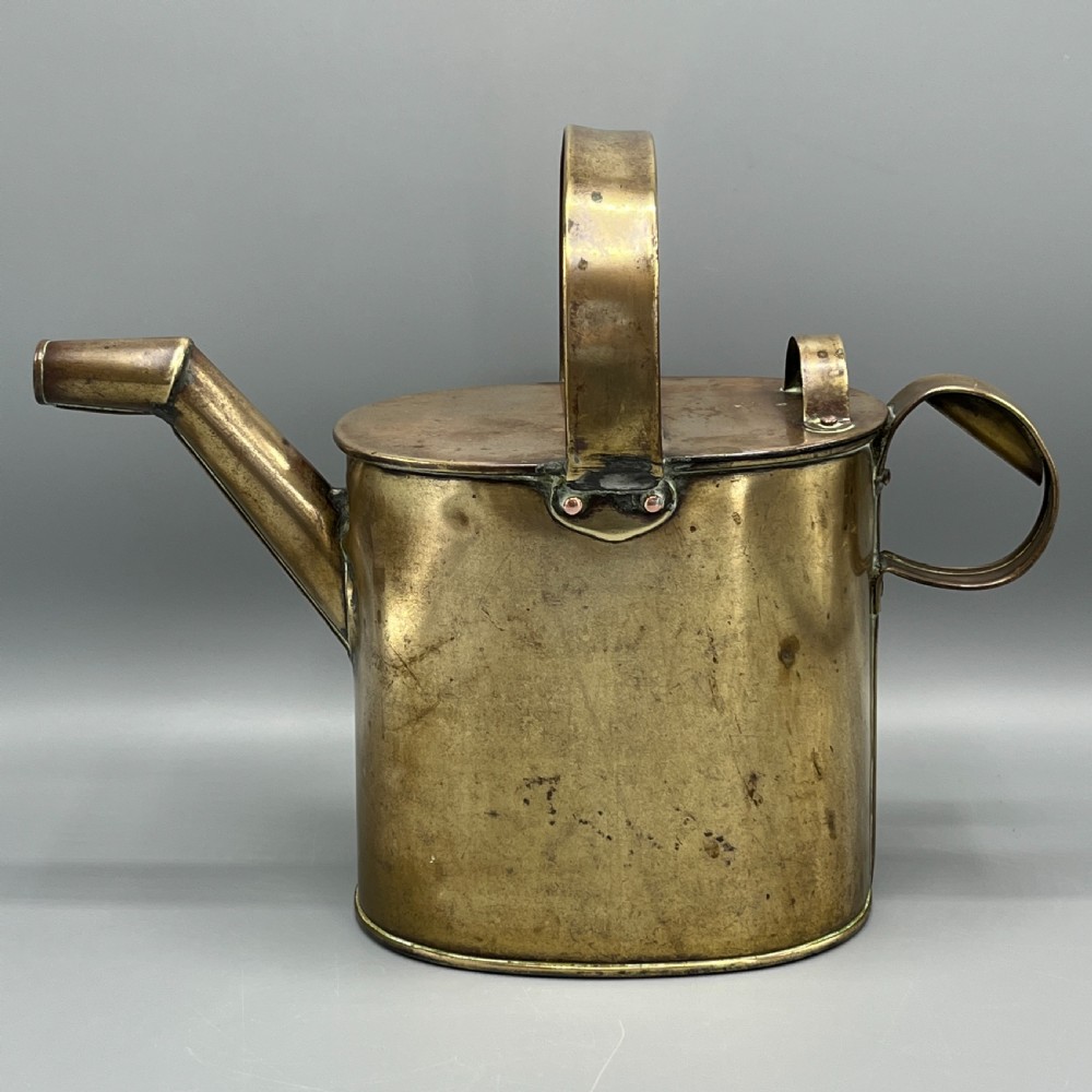 An Edwardian brass hot water or watering can featuring an oval body, fixed overhead flat strap handle, rear loop, diagonal spout, and visible copper rivets.