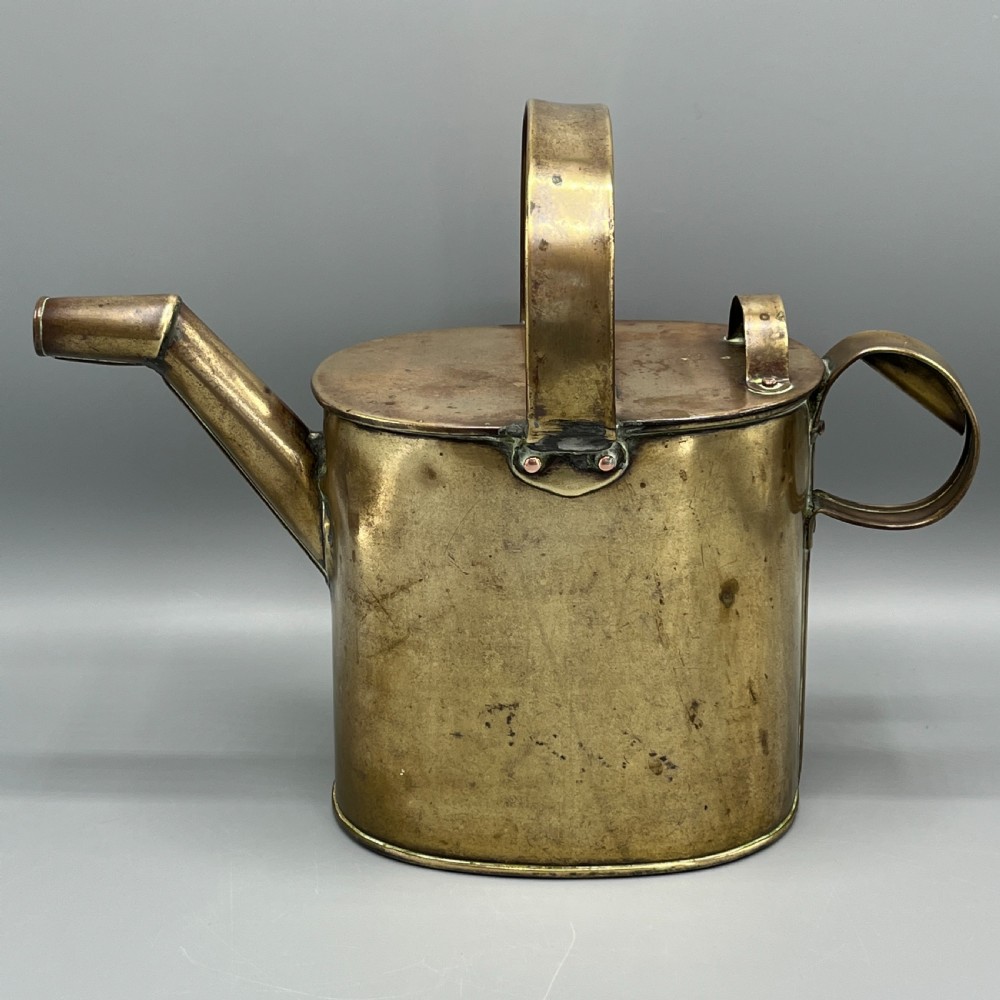 An Edwardian brass hot water or watering can featuring an oval body, fixed overhead flat strap handle, rear loop, diagonal spout, and visible copper rivets. View II