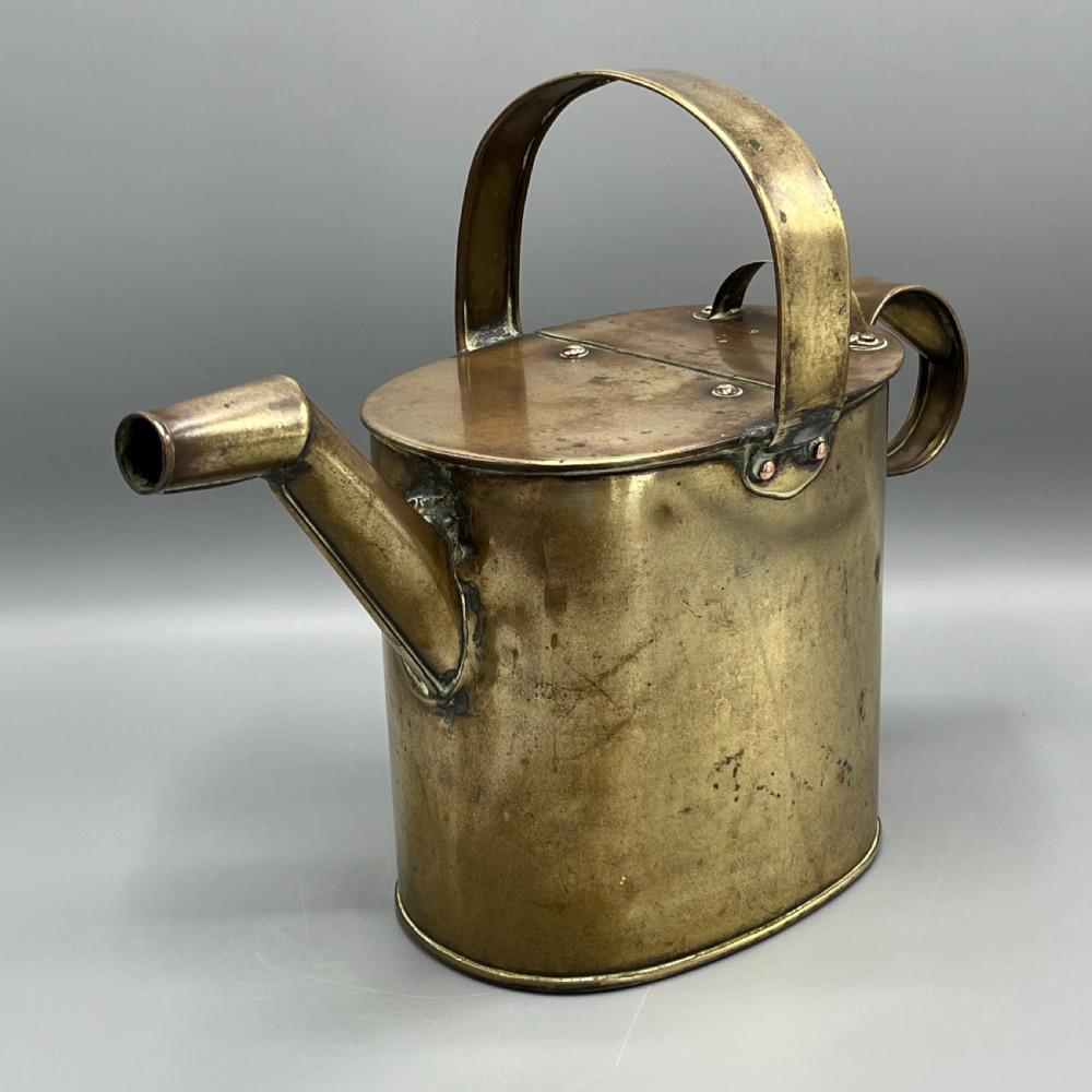 An Edwardian brass hot water or watering can featuring an oval body, fixed overhead flat strap handle, rear loop, diagonal spout, and visible copper rivets. View III
