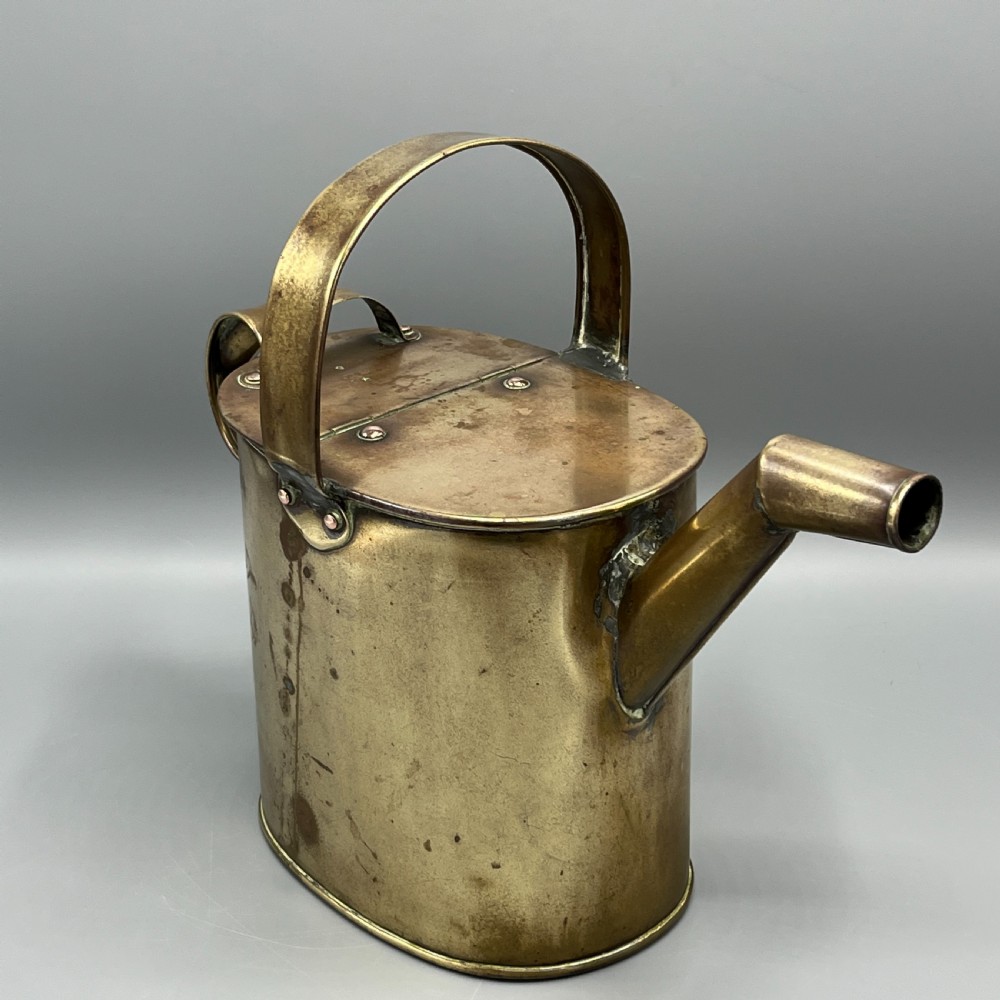 An Edwardian brass hot water or watering can featuring an oval body, fixed overhead flat strap handle, rear loop, diagonal spout, and visible copper rivets. View IV