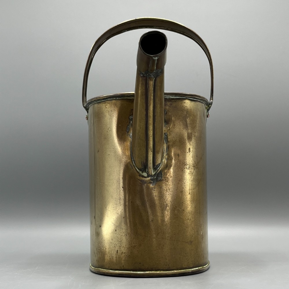 An Edwardian brass hot water or watering can featuring an oval body, fixed overhead flat strap handle, rear loop, diagonal spout, and visible copper rivets. View V