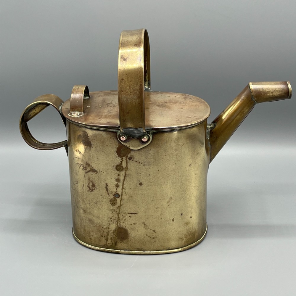 An Edwardian brass hot water or watering can featuring an oval body, fixed overhead flat strap handle, rear loop, diagonal spout, and visible copper rivets. View VI