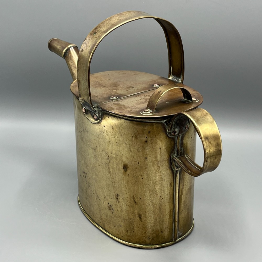 An Edwardian brass hot water or watering can featuring an oval body, fixed overhead flat strap handle, rear loop, diagonal spout, and visible copper rivets. View VII