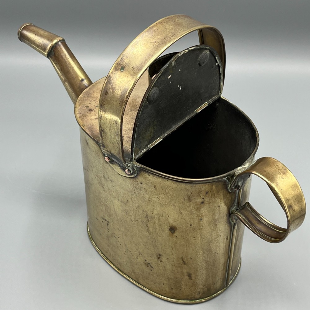An Edwardian brass hot water or watering can featuring an oval body, fixed overhead flat strap handle, rear loop, diagonal spout, and visible copper rivets. View IX