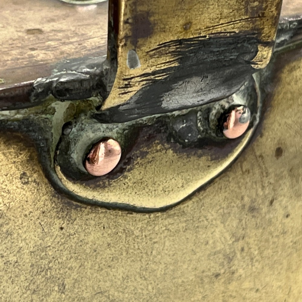An Edwardian brass hot water or watering can featuring an oval body, fixed overhead flat strap handle, rear loop, diagonal spout, and visible copper rivets. View X