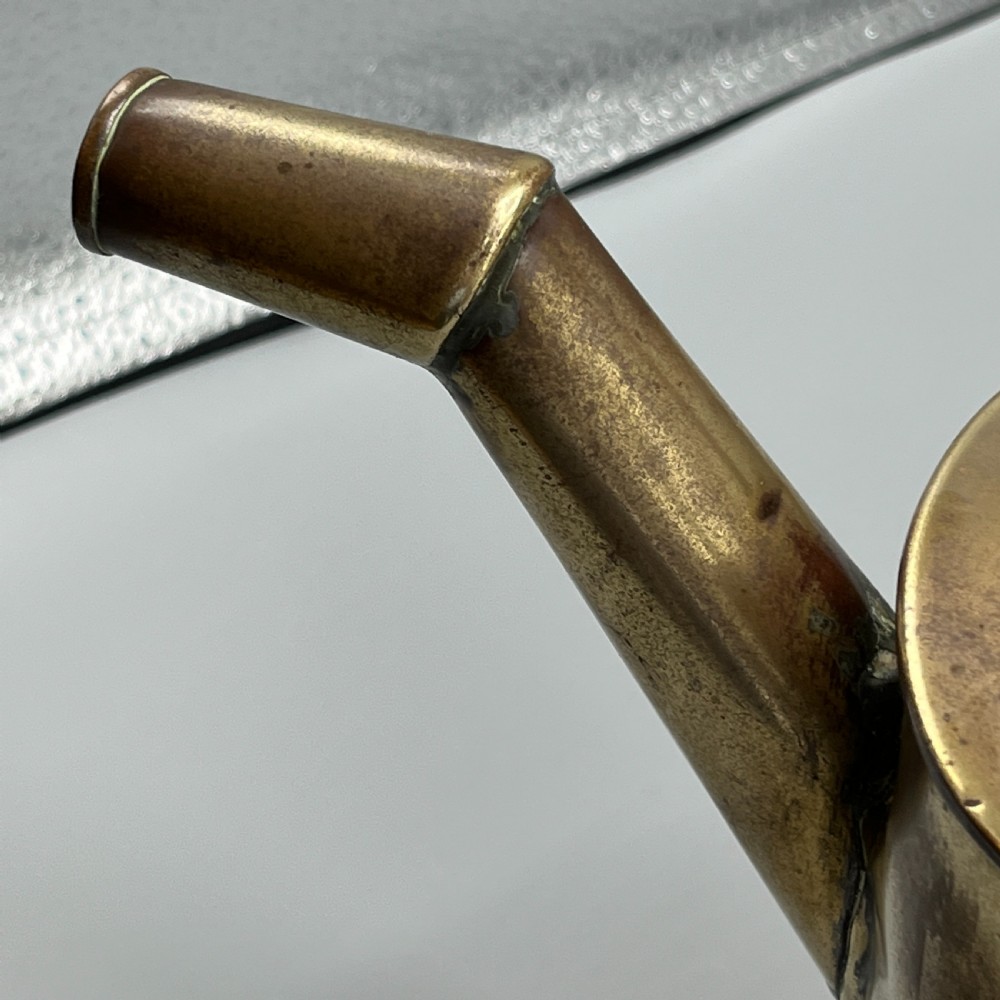 An Edwardian brass hot water or watering can featuring an oval body, fixed overhead flat strap handle, rear loop, diagonal spout, and visible copper rivets. View XI