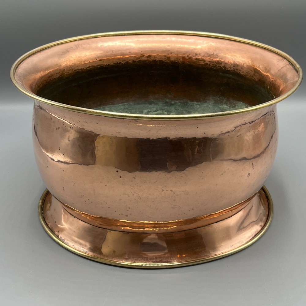 A large polished oval copper jardiniere with a flared rim edged in brass and a fitted flared brass base. View V