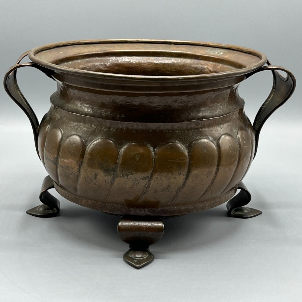 A hand-hammered solid copper Arts and Crafts jardiniere featuring curved side handles, lobed lower body detailing, and four raised feet.