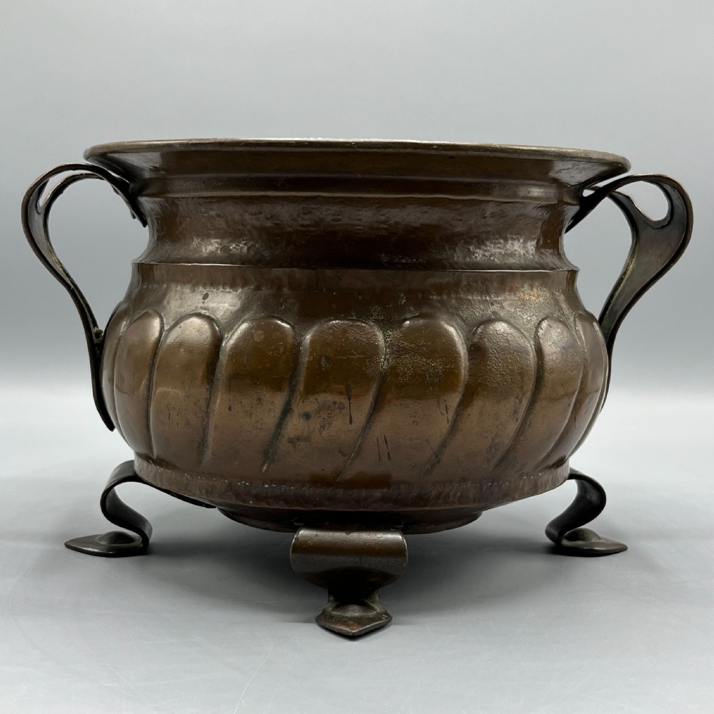 A hand-hammered solid copper Arts and Crafts jardiniere featuring curved side handles, lobed lower body detailing, and four raised feet. View III