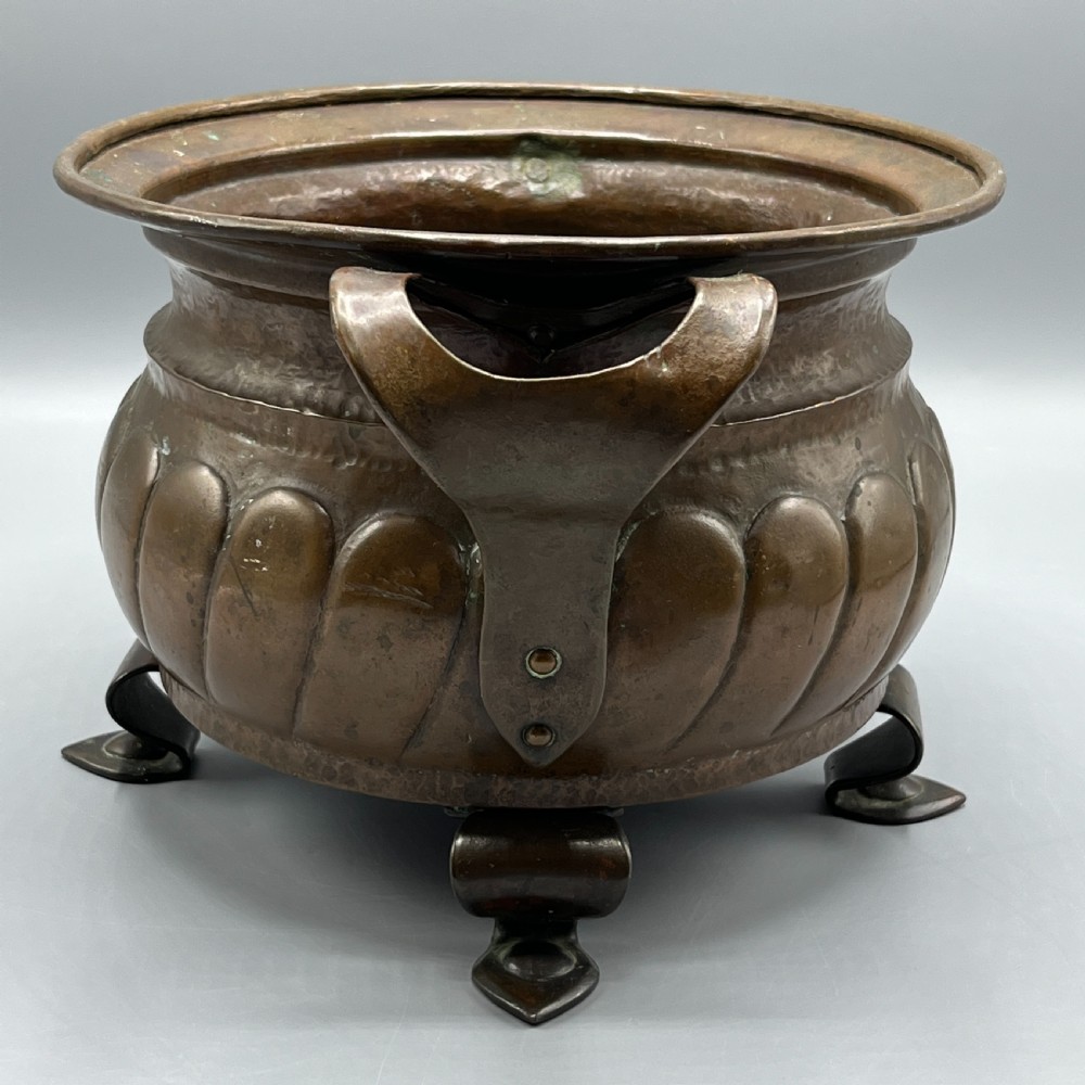A hand-hammered solid copper Arts and Crafts jardiniere featuring curved side handles, lobed lower body detailing, and four raised feet. View VI