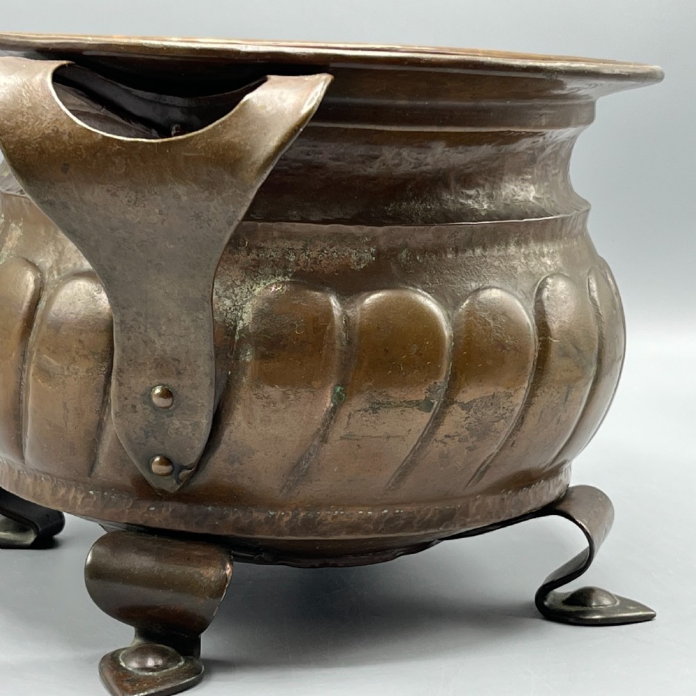 A hand-hammered solid copper Arts and Crafts jardiniere featuring curved side handles, lobed lower body detailing, and four raised feet. View X