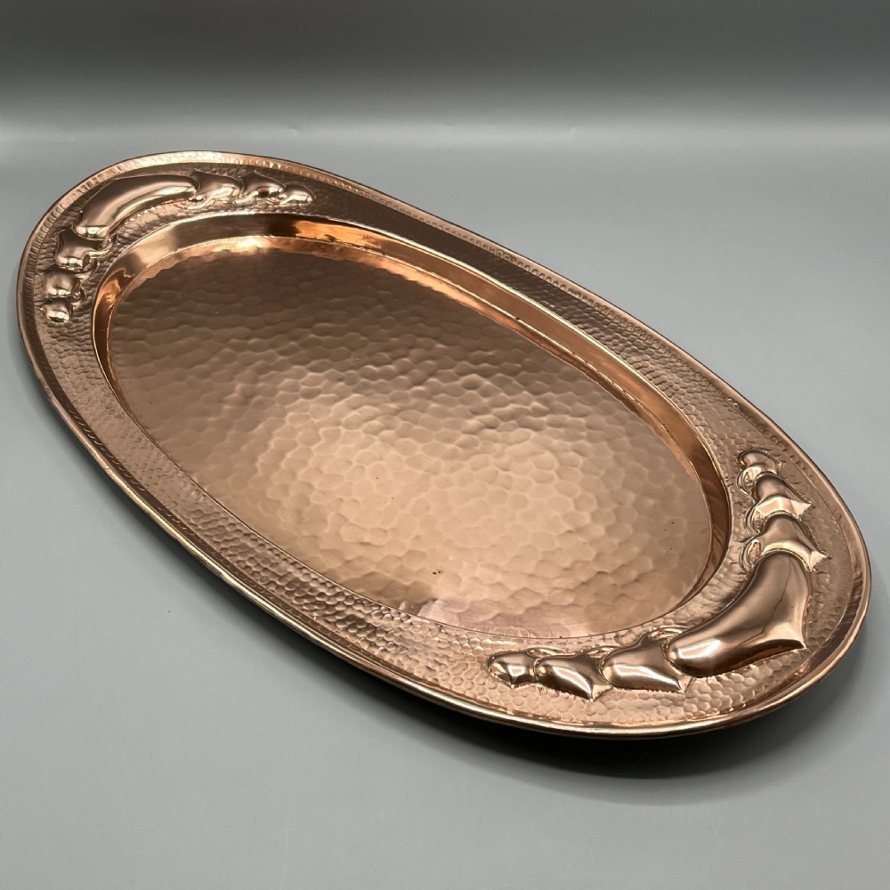 An oval Arts and Crafts copper tray featuring a planished hammered centre and stylised repousse detailing around the rim.