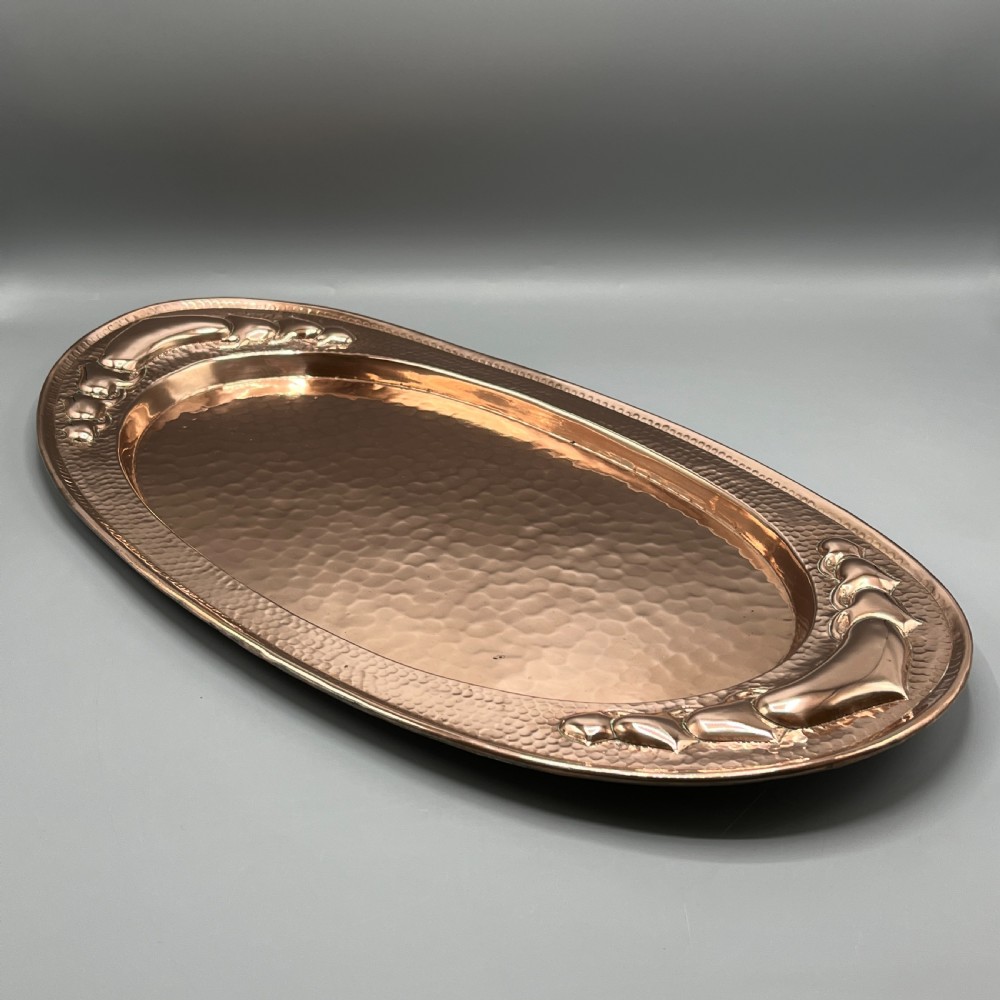 An oval Arts and Crafts copper tray featuring a planished hammered centre and stylised repousse detailing around the rim. View V