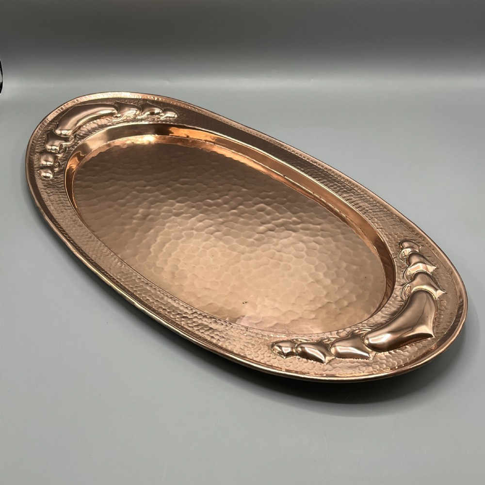 An oval Arts and Crafts copper tray featuring a planished hammered centre and stylised repousse detailing around the rim. View VI