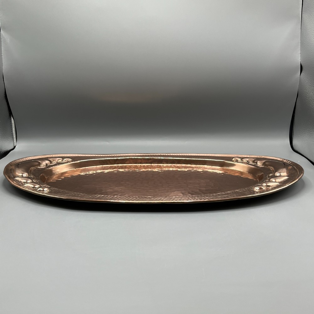 An oval Arts and Crafts copper tray featuring a planished hammered centre and stylised repousse detailing around the rim. View IX