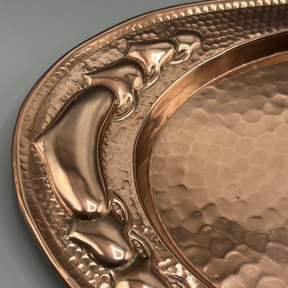 An oval Arts and Crafts copper tray featuring a planished hammered centre and stylised repousse detailing around the rim. View X