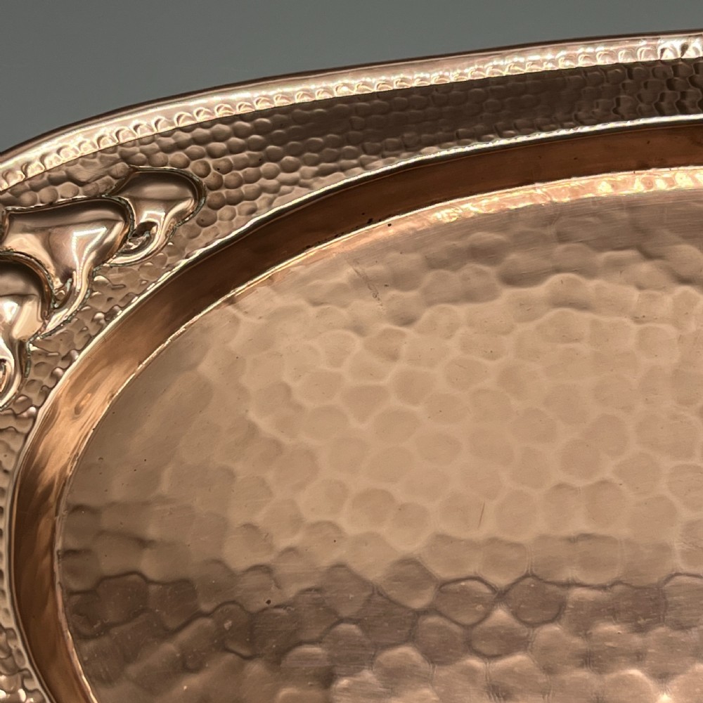 An oval Arts and Crafts copper tray featuring a planished hammered centre and stylised repousse detailing around the rim. View XII