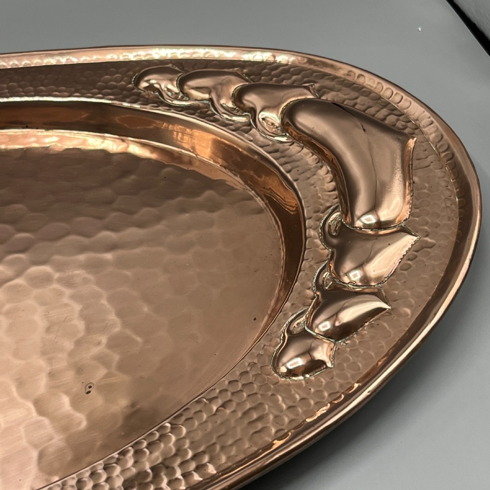 An oval Arts and Crafts copper tray featuring a planished hammered centre and stylised repousse detailing around the rim. View XVI