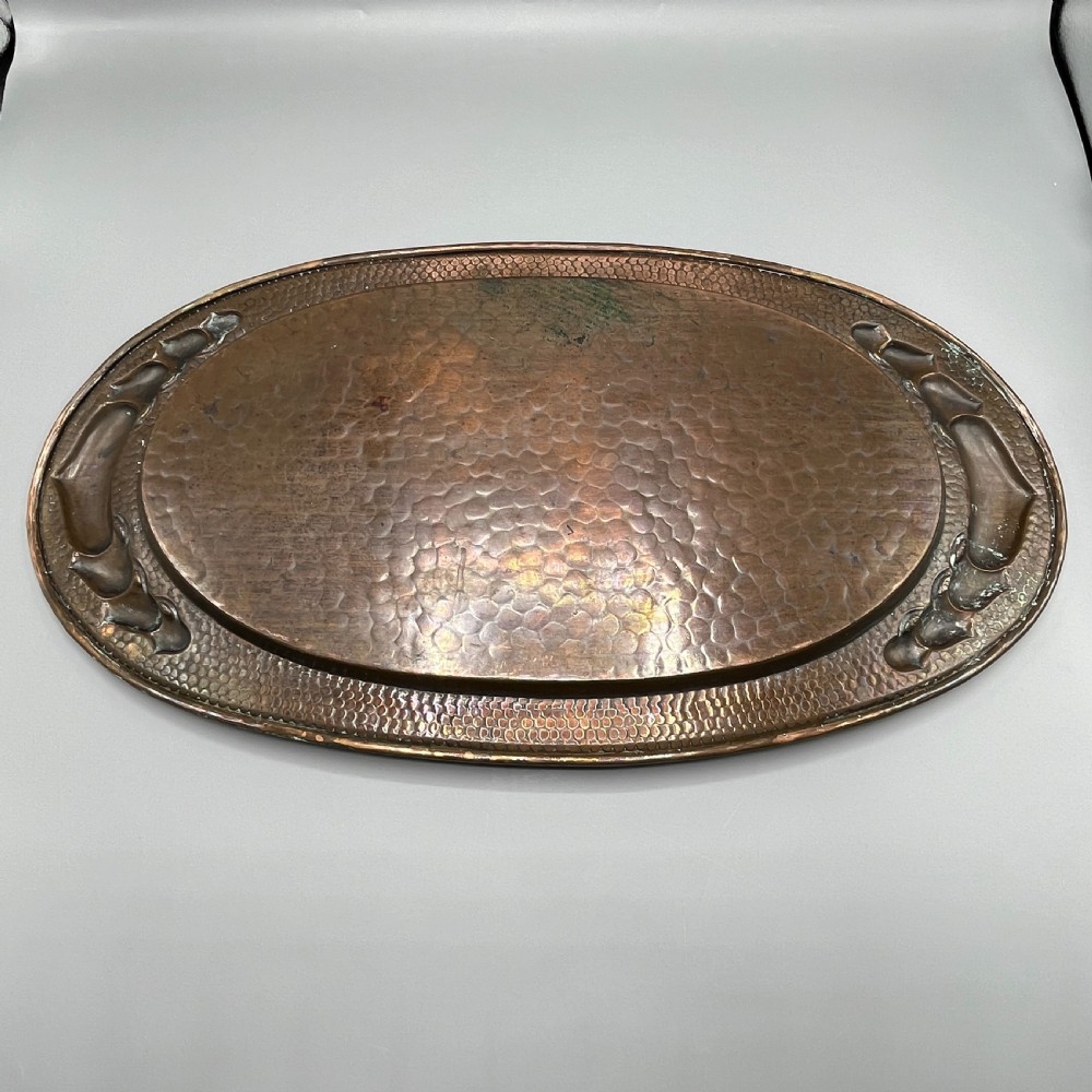 An oval Arts and Crafts copper tray featuring a planished hammered centre and stylised repousse detailing around the rim. View XX