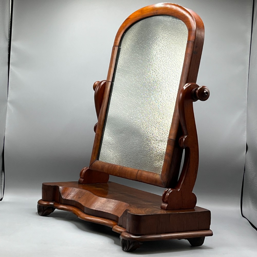 A Victorian mahogany dressing table mirror with an arched swivel frame and carved scroll uprights set on a serpentine base.